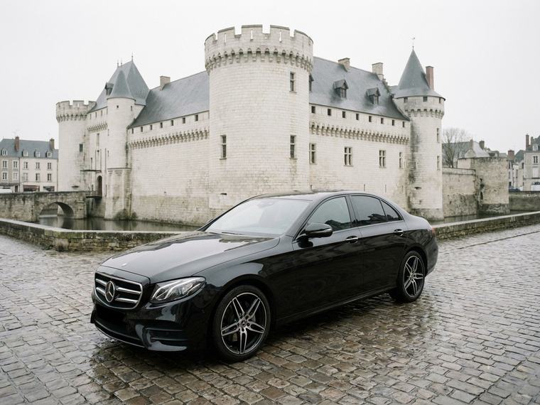 Berline noire sur une esplanade pavée devant un château aux remparts de pierre claire